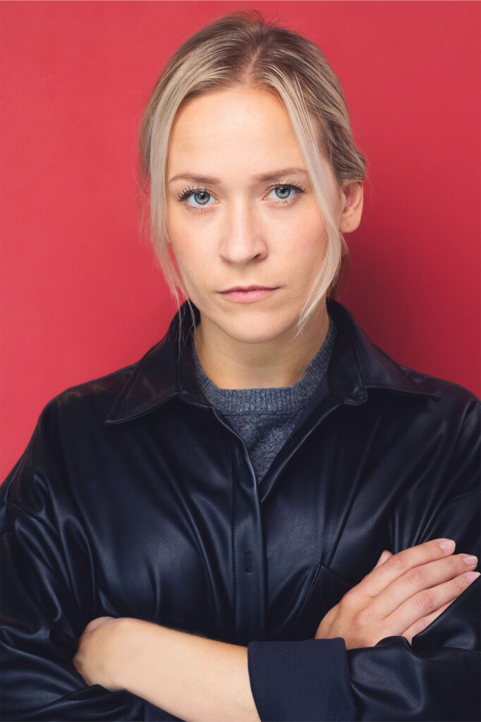 Actress, blonde with black leather jacket, crossing her arms, powerful, not approachable, red backdrop, nyc headshot backdrop