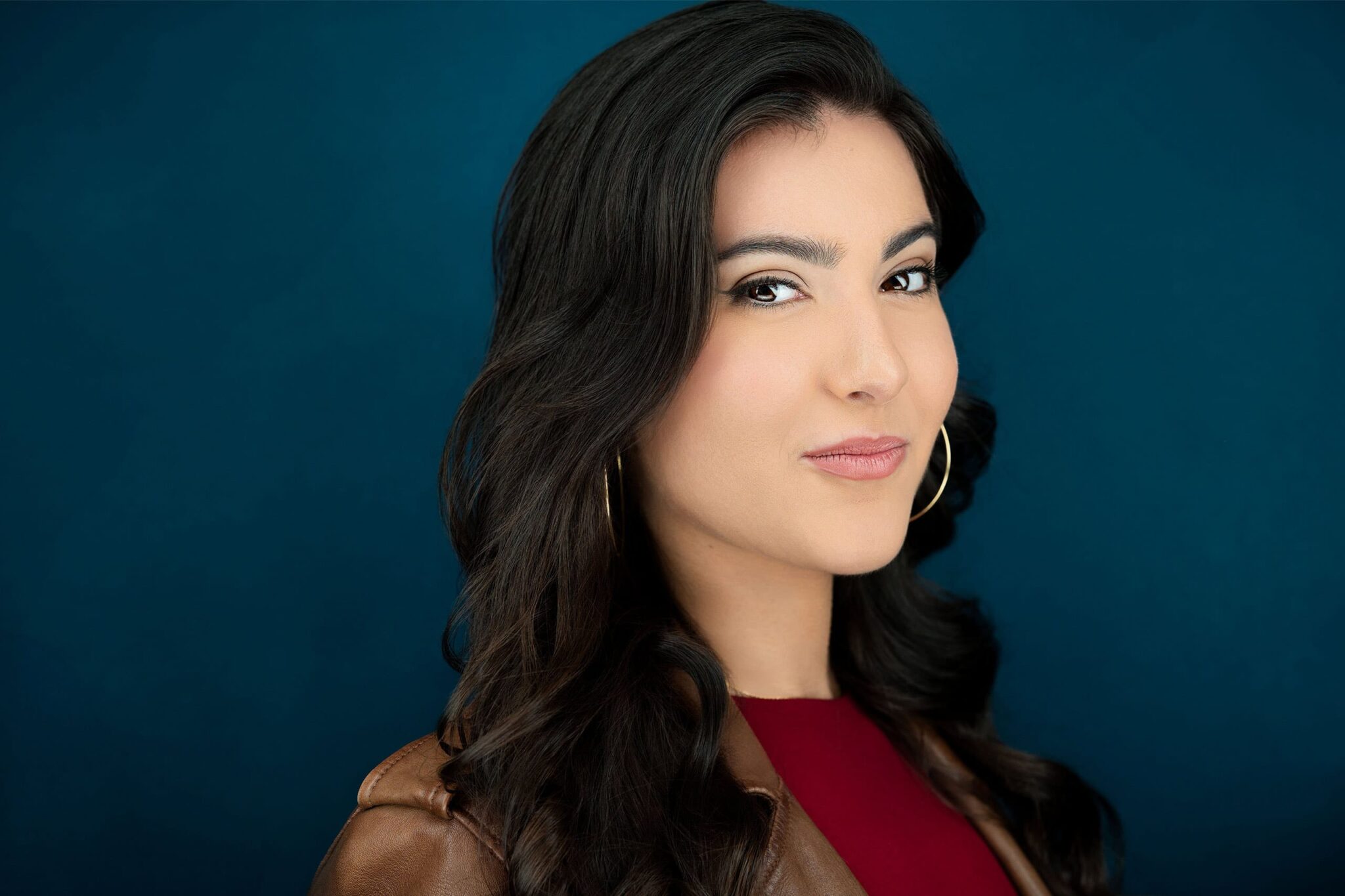 Stunning brunette actress with hoop earrings, confident smile, and inviting eyes, wearing a brown leather jacket and red shirt against a dark blue background. NYC Photographer
