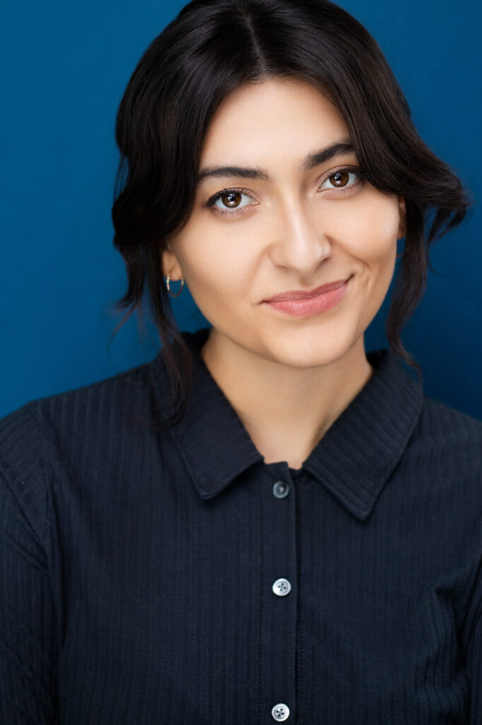 Brunette actress with hair up, strands of hair falling to the front of the face, button navy blue shirt, approachable, fun, great no teeth smile, blue backdrop, nyc headshot photographer