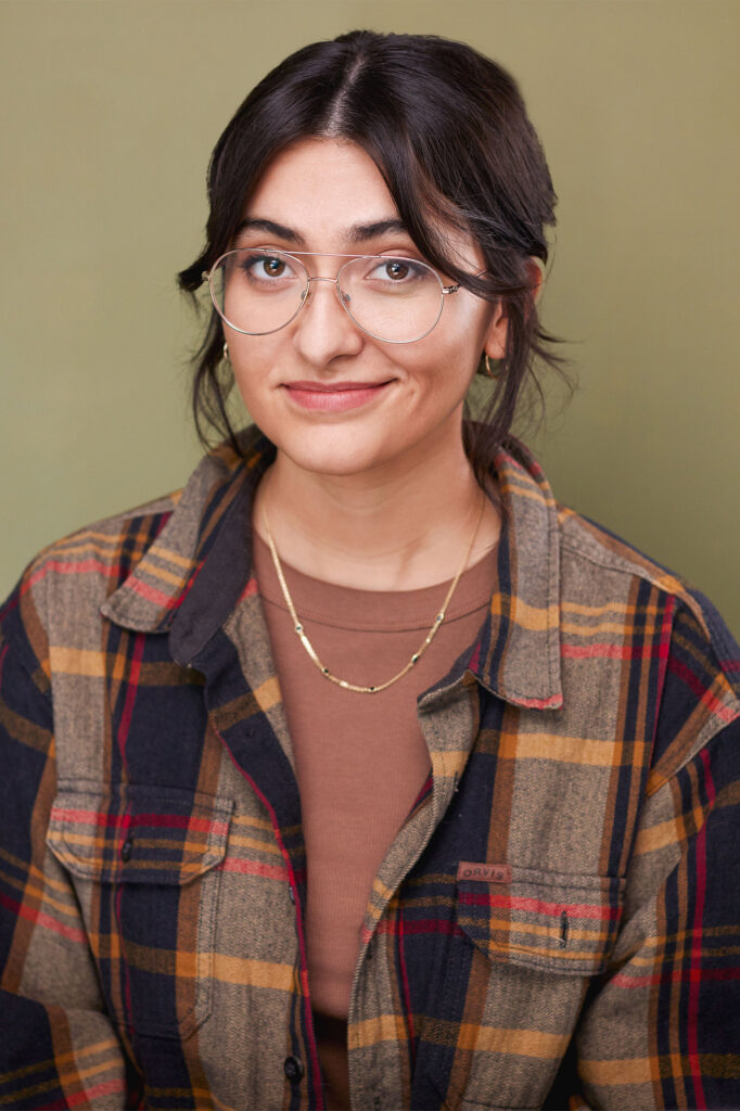 Actress with flannel, brown shirt, gold necklace, aviator type vision glasses, fun smirk, corky, olive green back drop, nyc headshot photographer