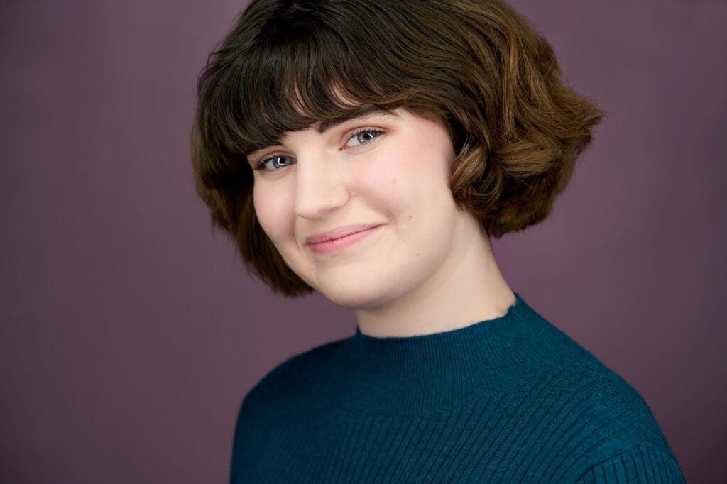 Brunette actress with approachable smile, purple backdrop, dark teal sweater, nyc photographer