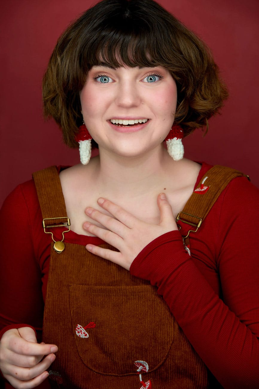 Fun portrait of an actress in brown corduroy overalls with mushroom patterns and a red shirt, giving a 'can't believe it' look with big blue eyes against a red background. NYC Headshot Photographer