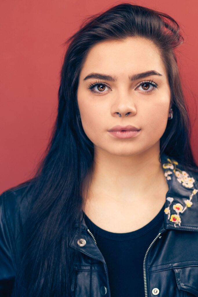 Actress with brown eyes, brown hair, nose ring, leather black jacket, black shirt, red background, strong powerful look, nyc headshot photographer