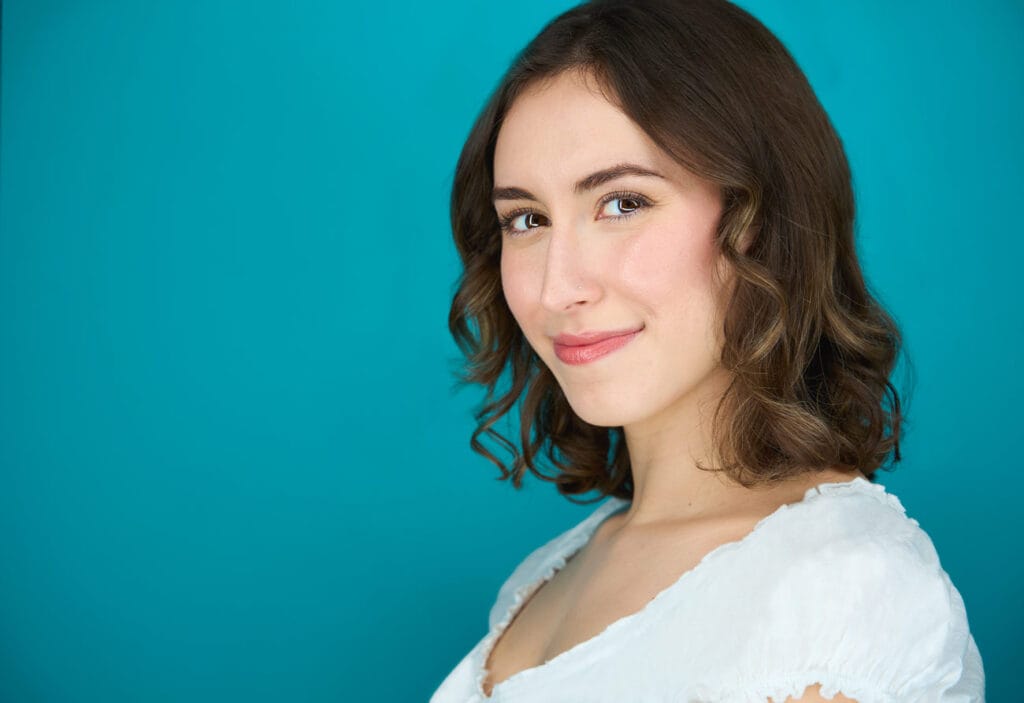 Actress turned profile but staring at camera, approachable smile, blue backdrop, white blouse, actress, headshot, nyc photographer