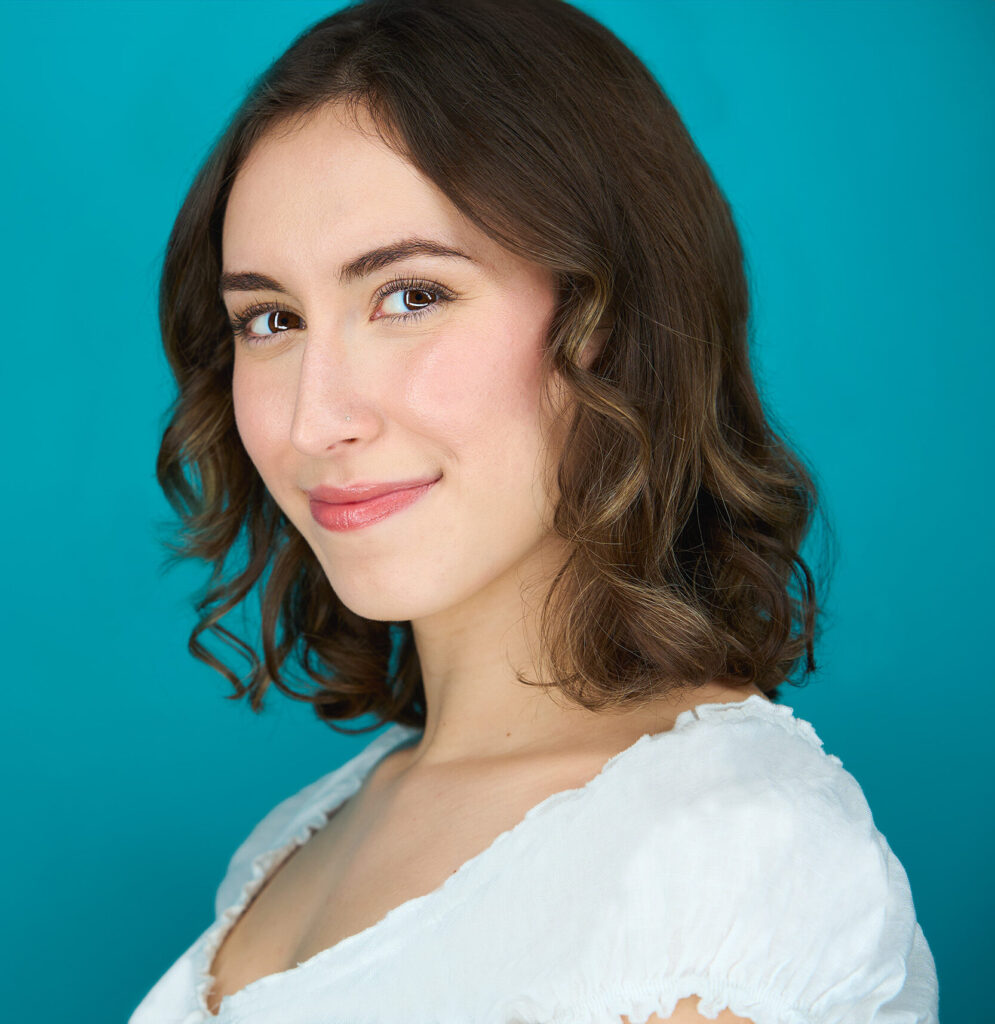 Actress turned profile but staring at camera, approachable smile, blue backdrop, white blouse, actress, headshot, nyc photographer