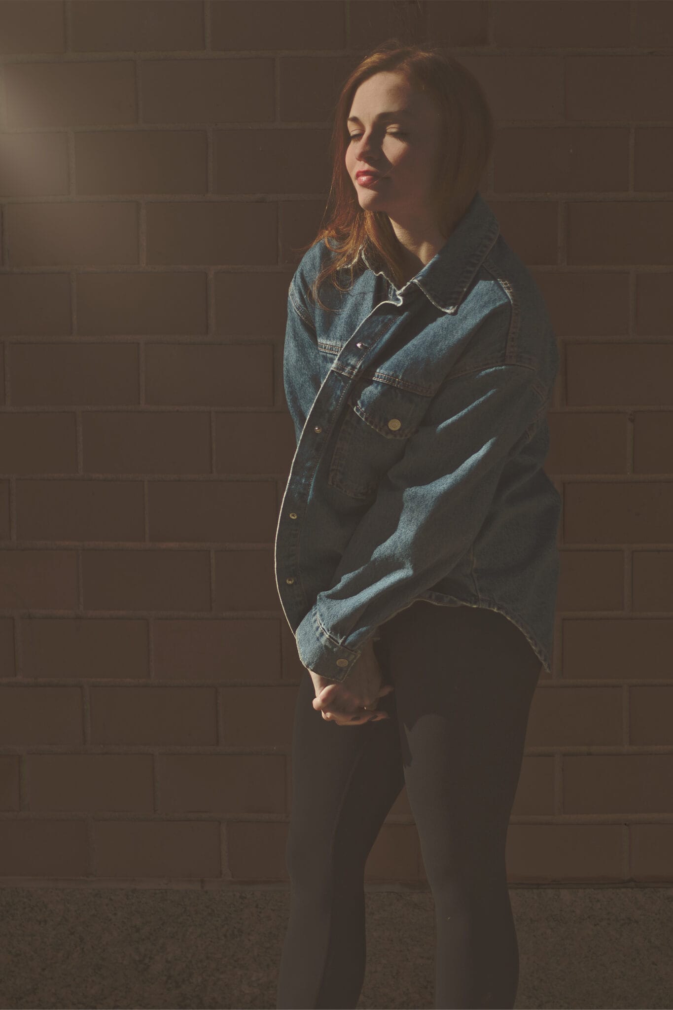 Redhead with closed eyes, basking in the sun, pulling her jean jacket close in the cold. Serene, meditative moment against a brick wall backdrop. Portrait by NYC photographer.