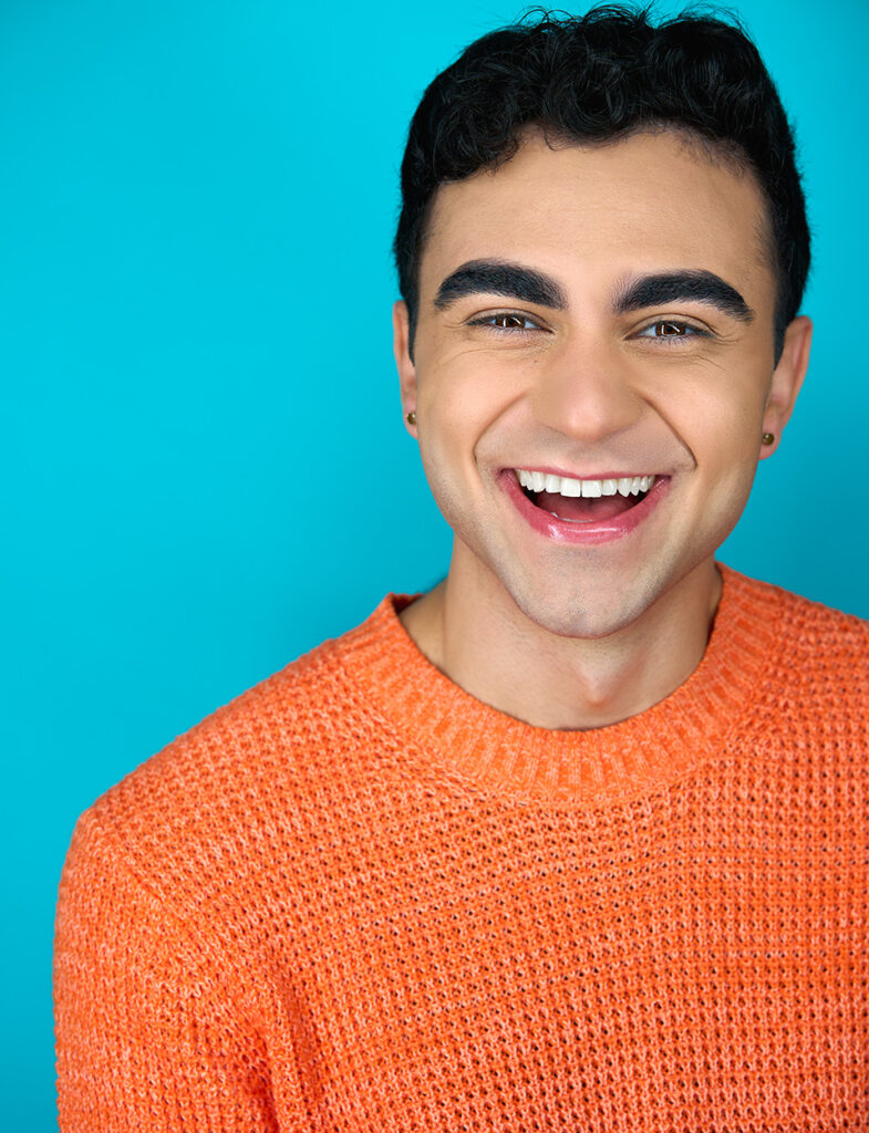 Male actor smiling/half laugh towards the camera, orange sweater, teal background, headshot, actor, nyc photographer