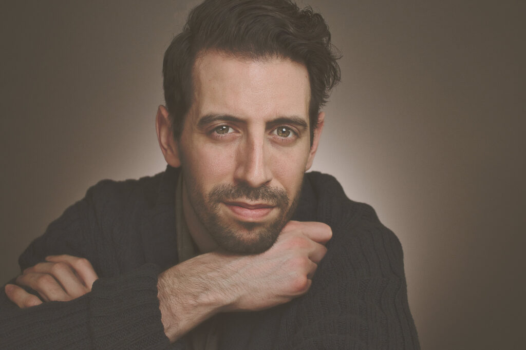 Self Portrait of photographer, arms folded, leaning on table top, gazing at the camera, approachable smile, vaseline backdrop, hazel eyes, nyc photographer