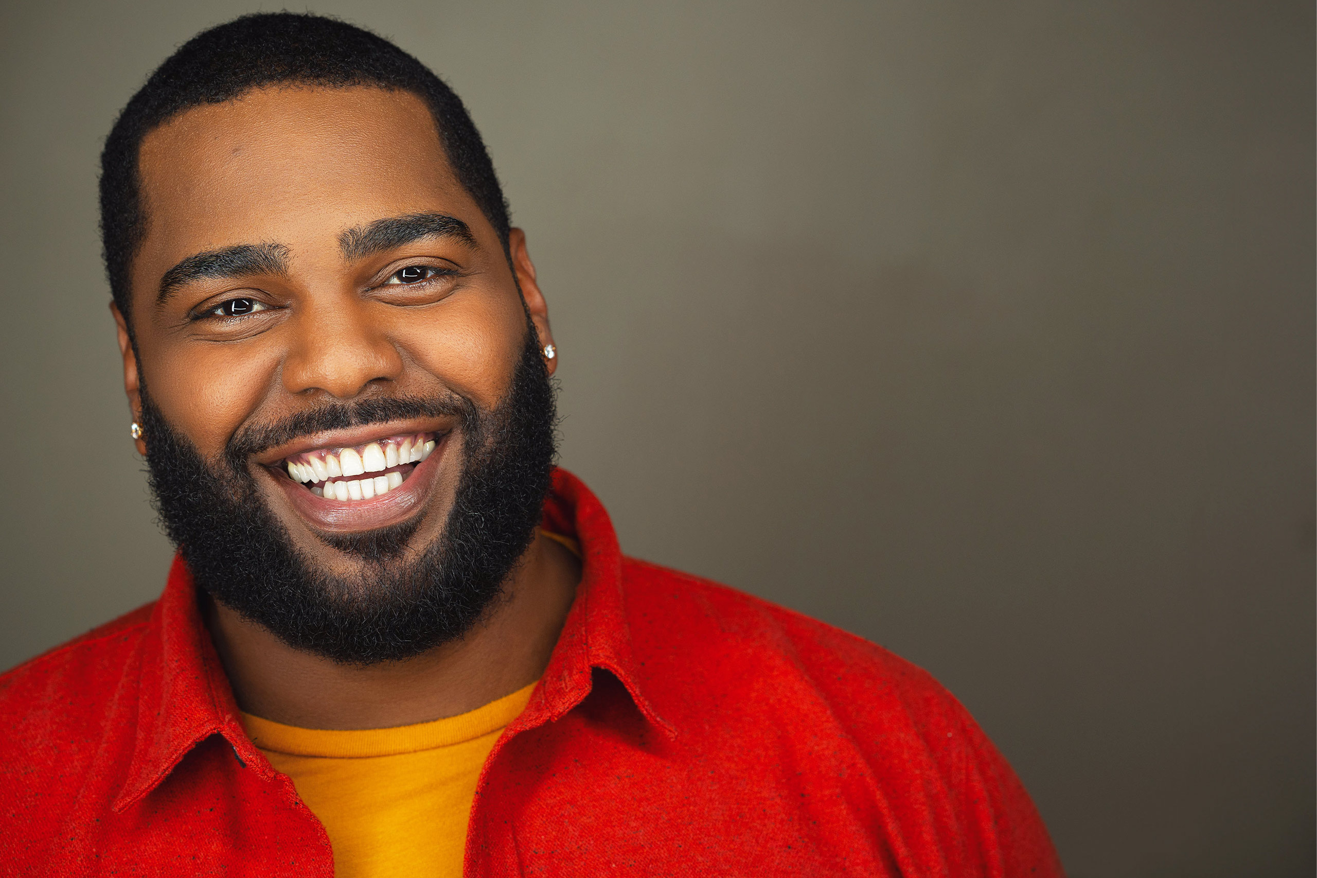 Black actor in a headshot with a beard and a big, bright smile, wearing an orange button-down over a yellow shirt, in front of an olive green background. NYC Headshot Photographer