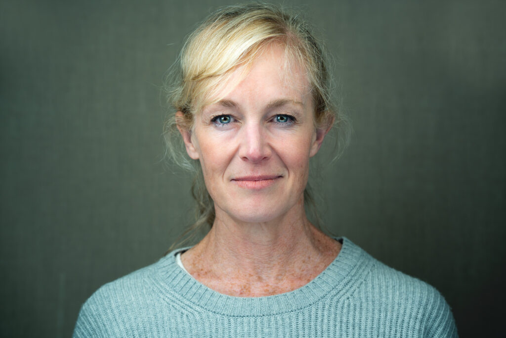 Raw photo of a woman squared to the camera, staring confidently with blue eyes and messy hair, against a textured beige backdrop. NYC Headshot Photographer