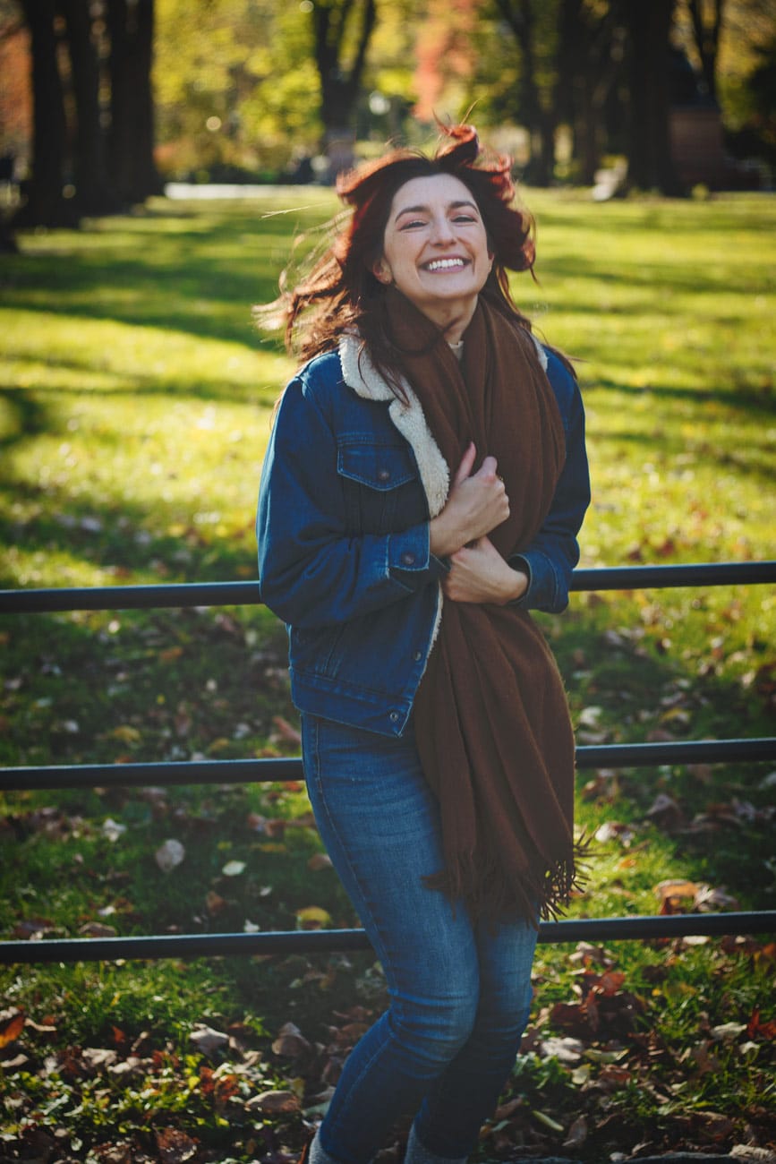 Candid portrait of a model with a joyful smile, jumping in Central Park during fall. Her hair is flying upward as she holds her scarf and wears a jean jacket over her outfit. Vibrant scene with green trees in the background. NYC photographer.