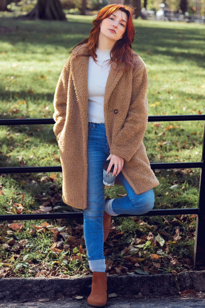 Model in Central Park holding a coffee cup, wearing a long brown furry jacket with jeans, red hair, and an editorial, carefree expression.
