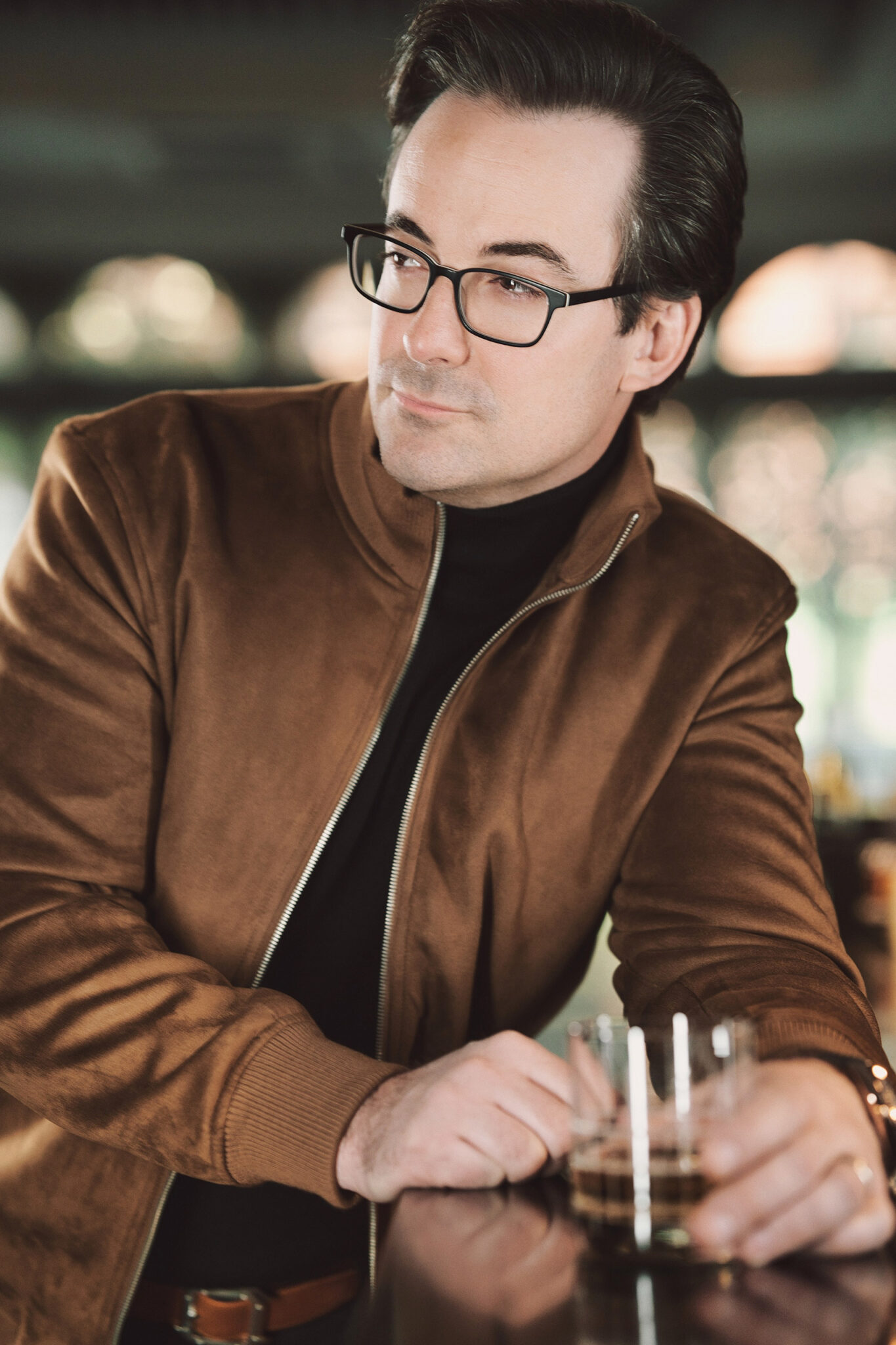 Gentleman in a lifestyle portrait leaning on a bar with a cocktail in hand, gazing out and away from the camera, exuding a sharp and upscale vibe. NYC Photography, portrait