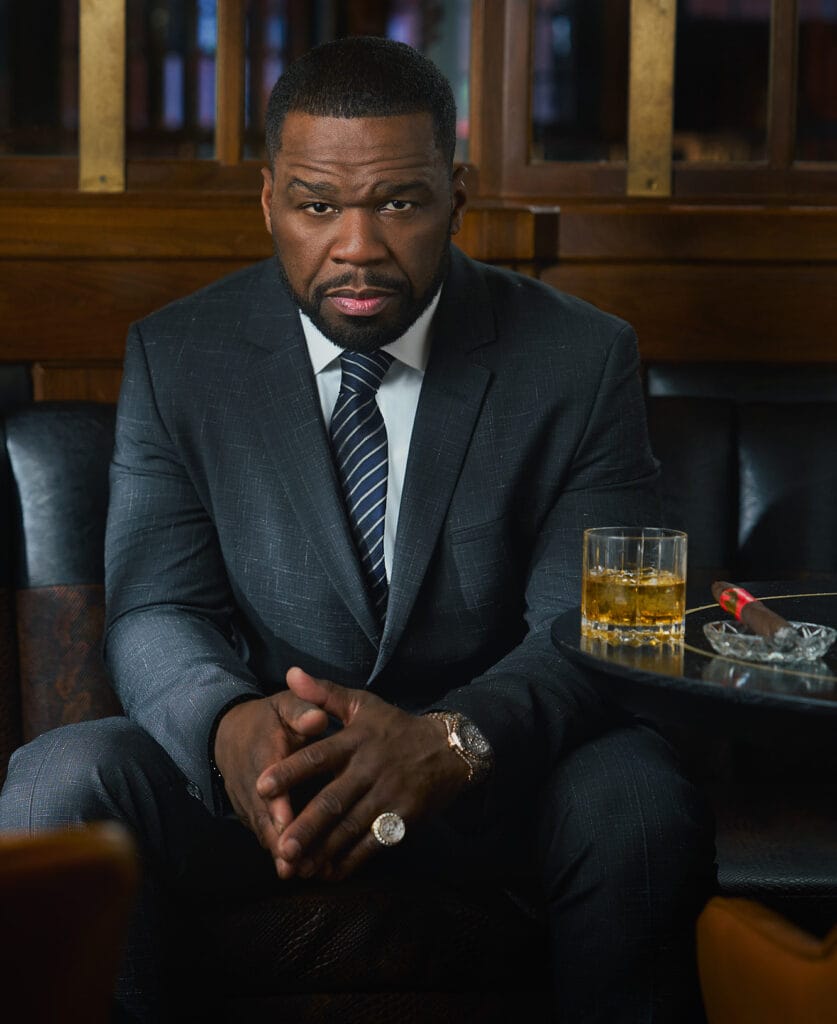 50 Cent sitting on banquet staring at camera, stern, business like gaze, glass of cognac on the table with lit cigar. NYC Photographer