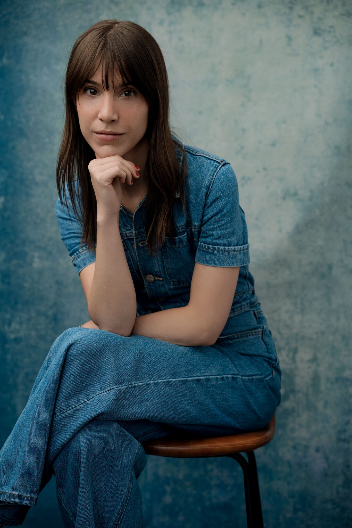 NYC Editorial shot of woman, jean jumpsuit, crossed legs with crossed arms leaning over gazing at the camera, one hand under chin - thinking look, blue jean like backdrop, nyc photographer, lifestyle, portrait Photography