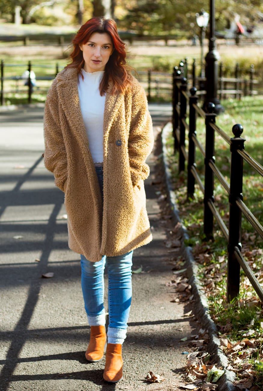 Woman in jeans and a brown overcoat walking directly toward the camera in Central Park during fall.