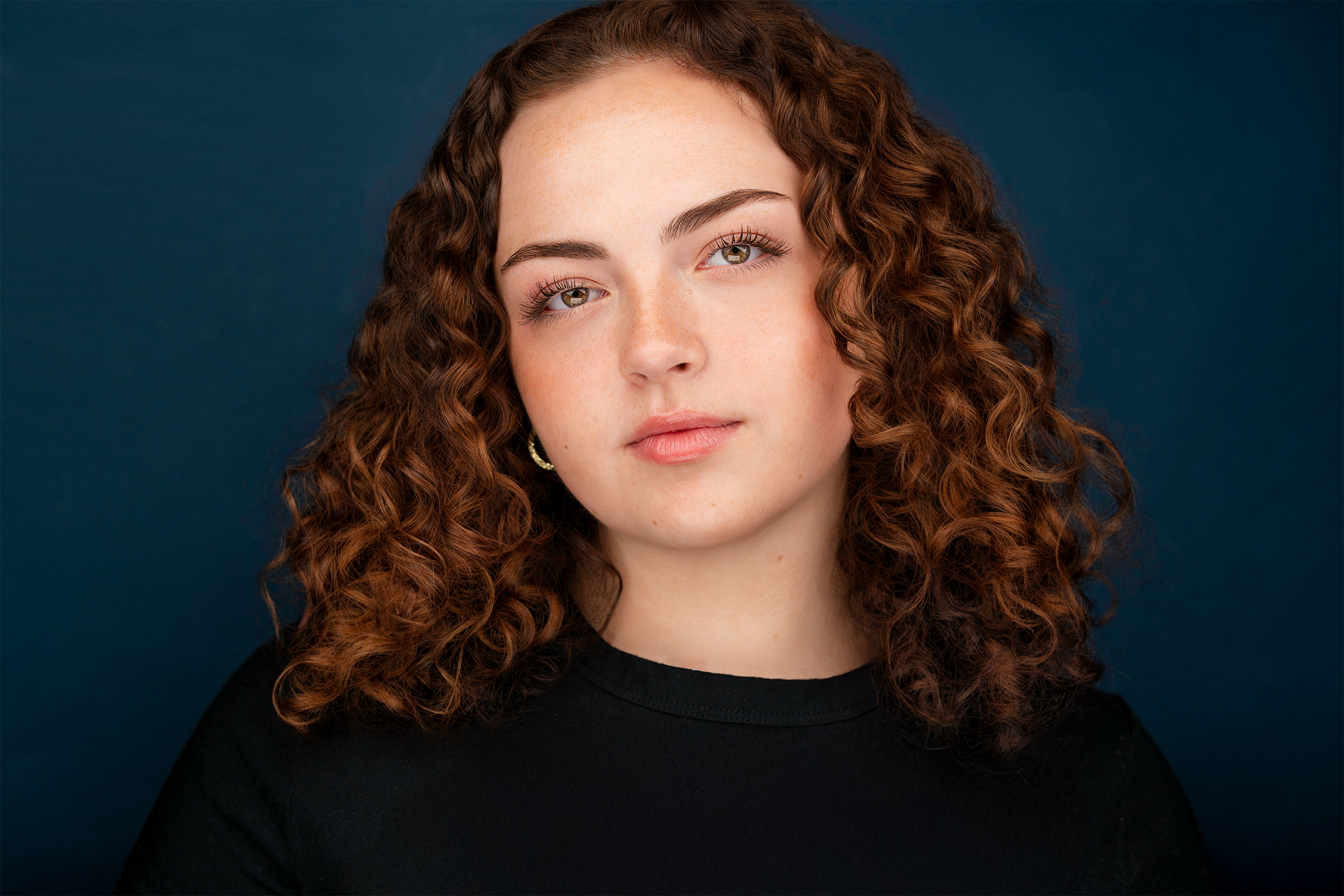 Young lady with curly brown hair, black top, blue backdrop, actress, headshot, drama, nyc