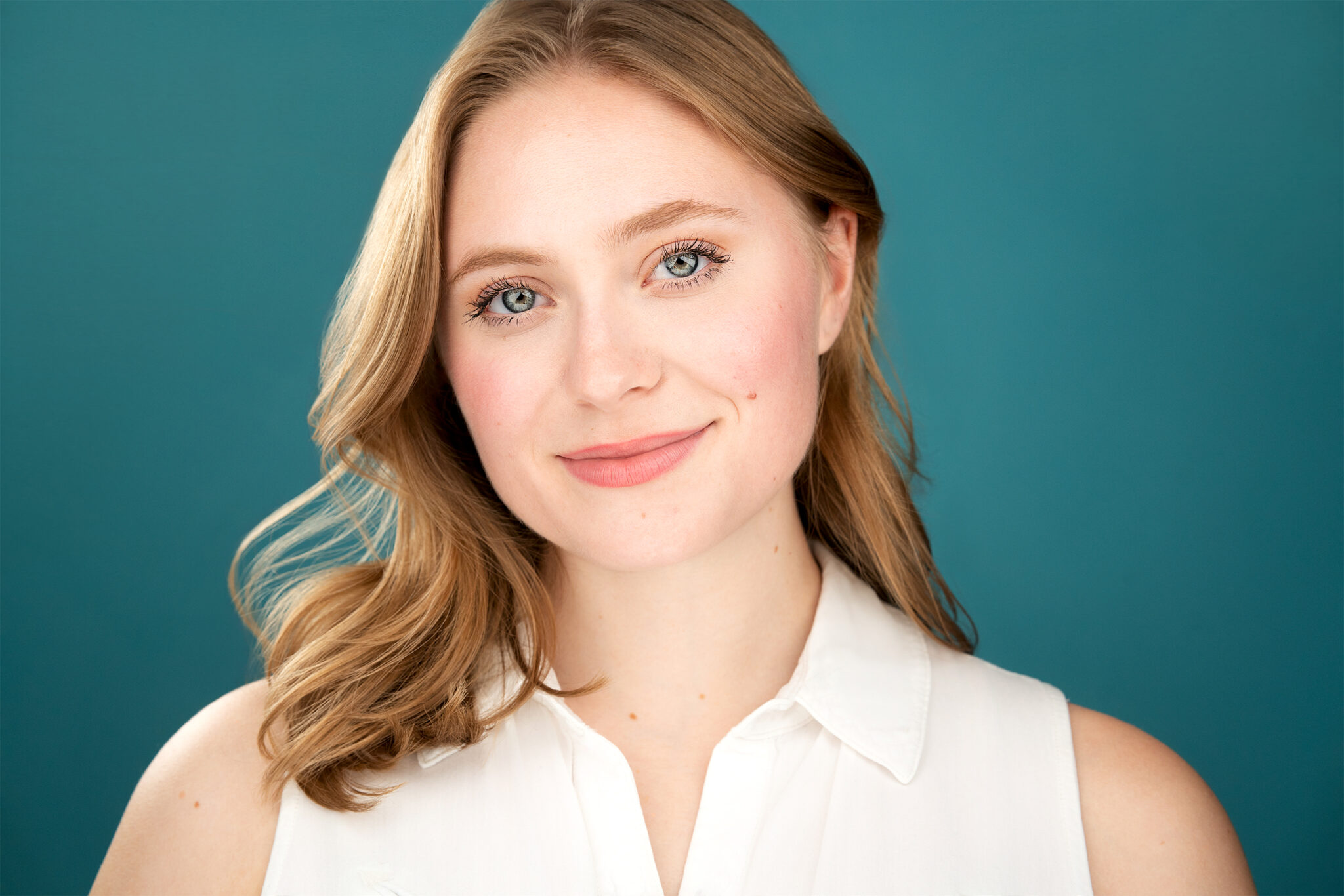 Approachable smile from a young lady with strawberry blonde hair, white collared sleeveless shirt, bright blue backdrop, warm, fuzzy, actress, headshot, nyc