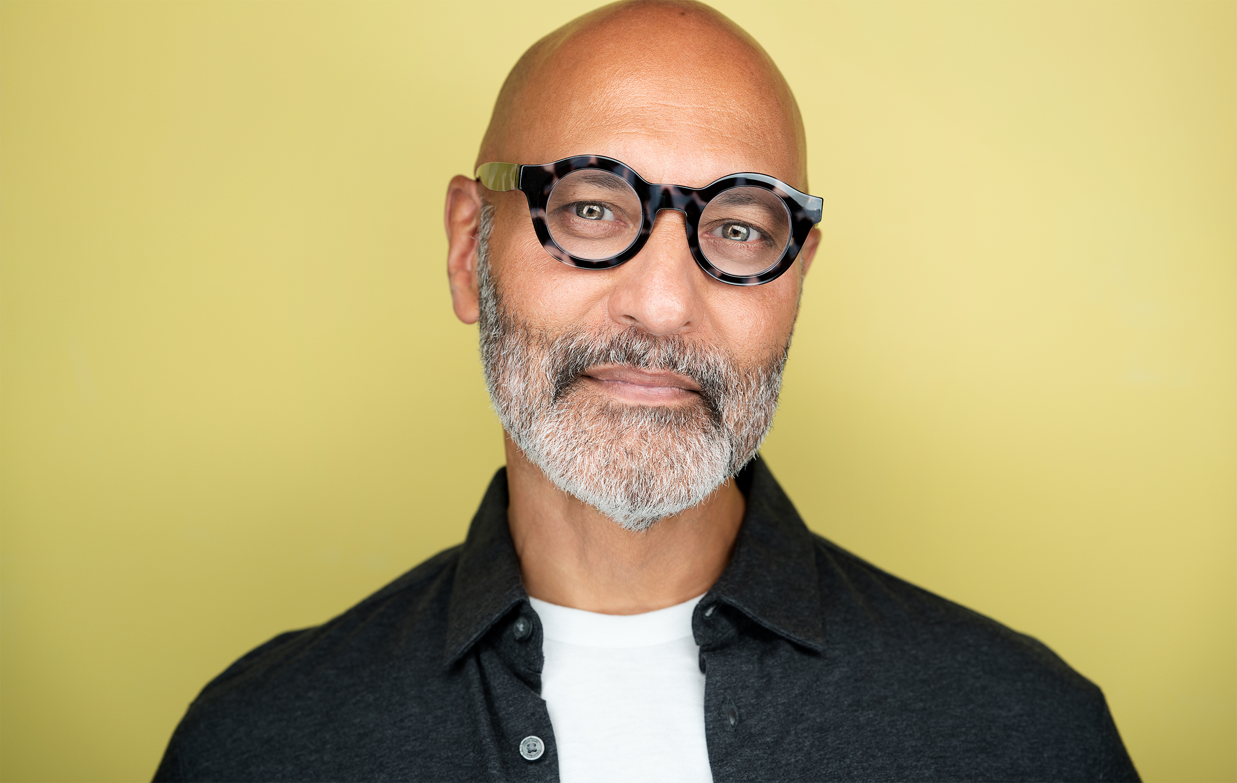 Bald man with glasses, gray beard, unbuttoned gray shirt and white shirt underneath, staring at camera, yellow backdrop, fun, actor, headshot, nyc