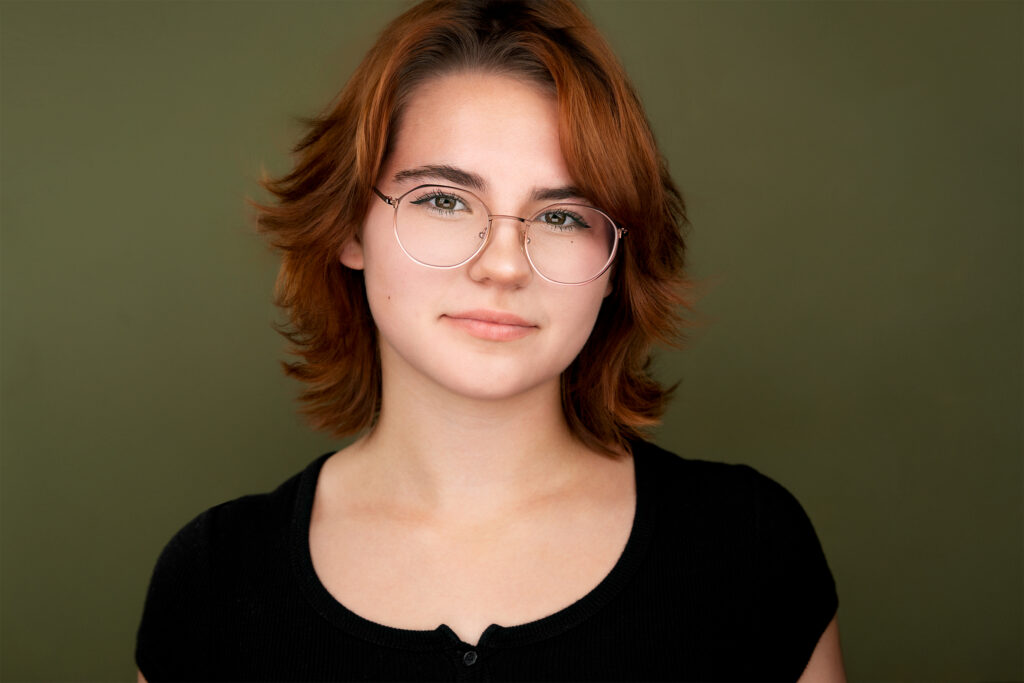 Woman wit glasses, red hair, black top, green backdrop, gazing at the camera, actress, headshot, NYC