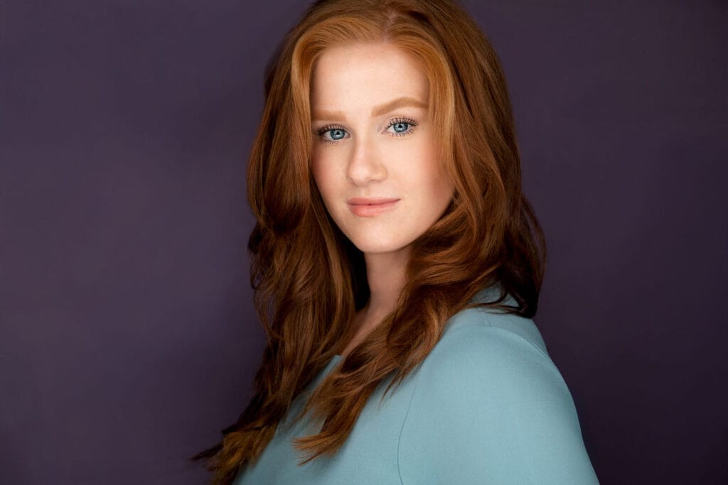 Woman with blue eyes and red hair looking at camera, headshot, light blue top and purple backdrop