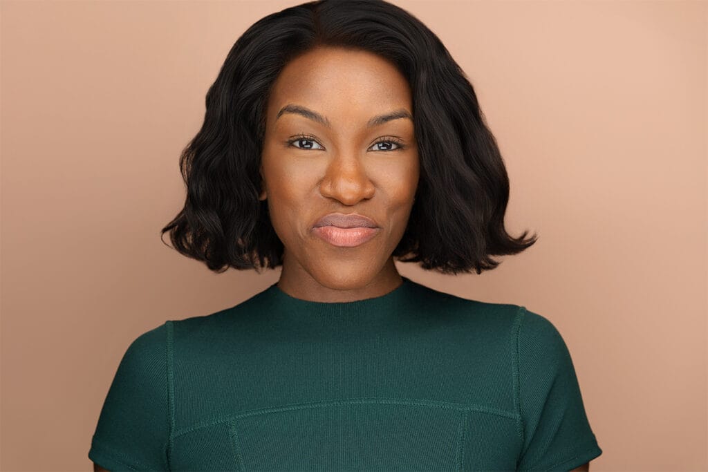 Woman of color with a nice smize, peach background, deep earthy green shirt, black hair, headshot