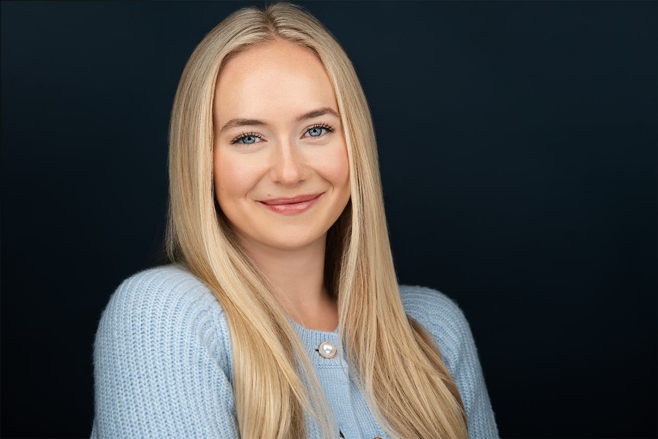 Blonde, Blue eye young lady, baby blue cardigan, dark blue backdrop, smiling no teeth to the camera. NYC Headshot Photographer