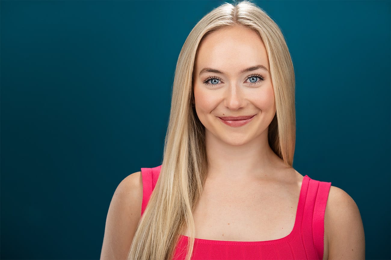 Blonde, Blue eye young lady, smiling no teeth to the camera straight on, pink top, teal backdrop