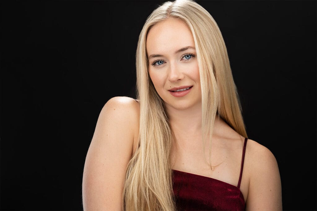 Blonde, blue eye young lady, seductive look, one shoulder hiked up, sensual, black backdrop, velvet red top. NYC Headshot Backdrop