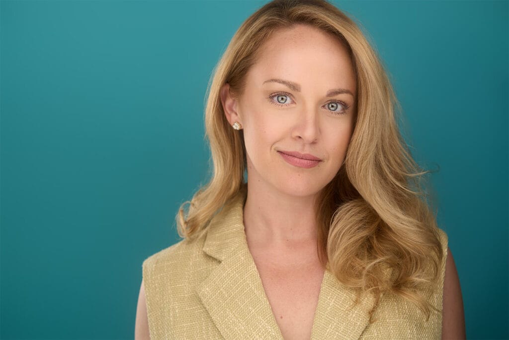 Blonde, blue eye lady, with beige top, teal backdrop, sly smile, approachable, NYC Photographer
