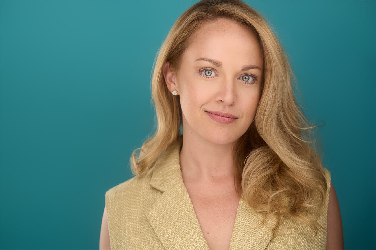 Blonde, blue eye lady, with beige top, teal backdrop, sly smile, approachable, NYC Photographer