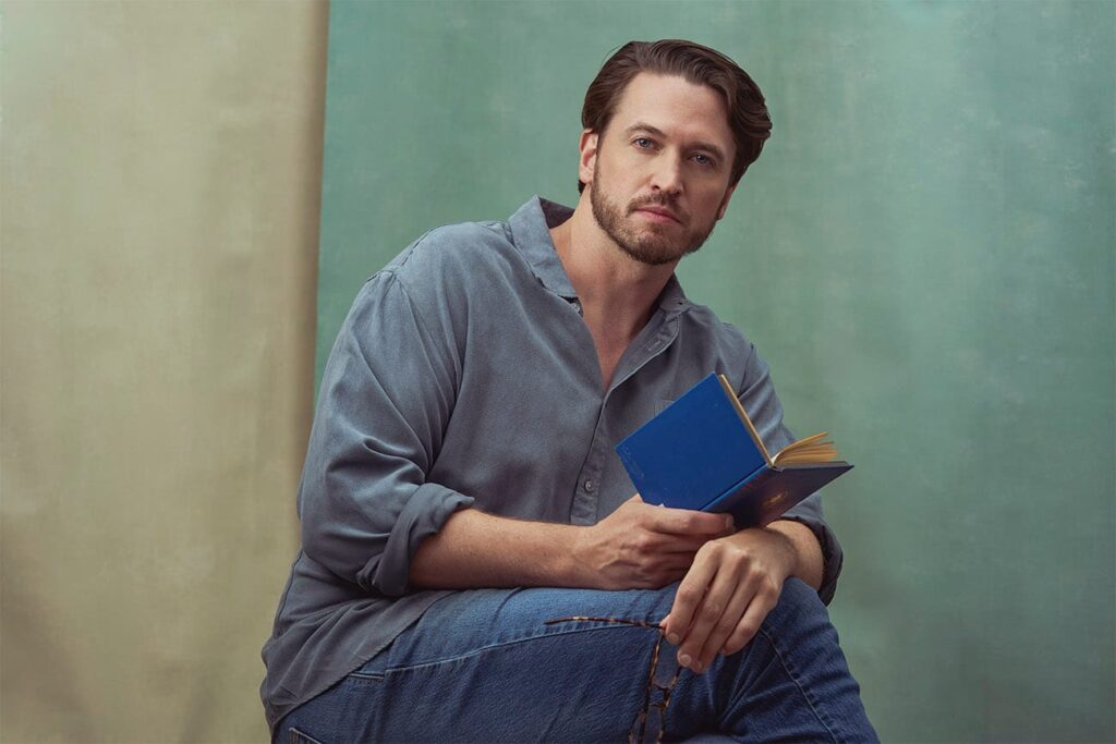 Gentleman with denim like button down shirt, denim jeans, sitting holding a poetry book and glasses staring at the camera, faded, muted green and beige backdrops, portrait, nyc photographer