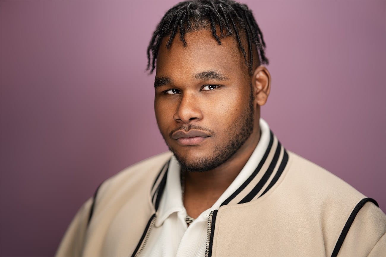 Young man, black hair, semi beard, piercing gaze, champagne jacket with white polo, purple backdrop, nyc headshot photographer