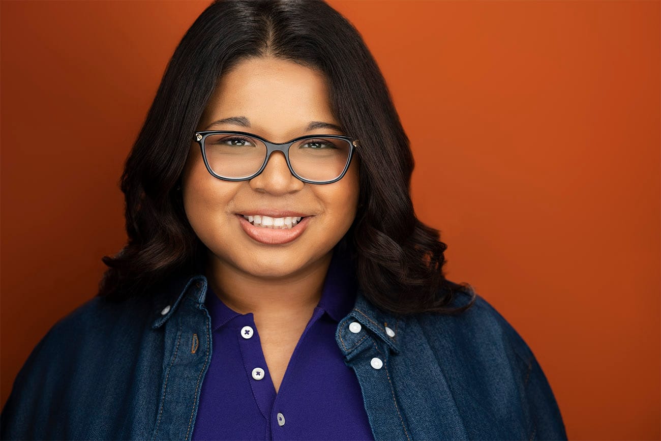 Young Lady with glasses, smile, purple polo with denim button down layered, orange backdrop, brown hair, nyc photographer