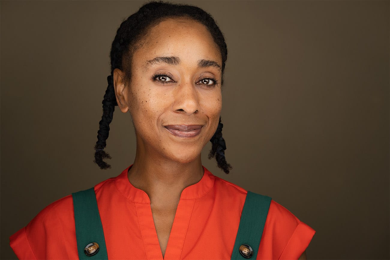 Approachable woman with closed mouth smile, braids, freckles, orange top with green overalls, headshot, muted earthy color backdrop, nyc photographer