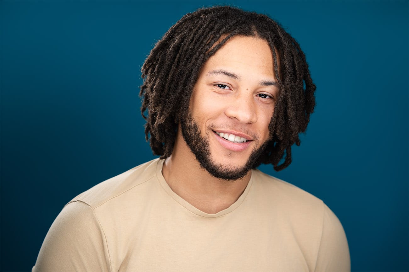 Young gentleman in beige top, dark blue backdrop, soft open smile, beard, short dreads, actor, nyc photographer